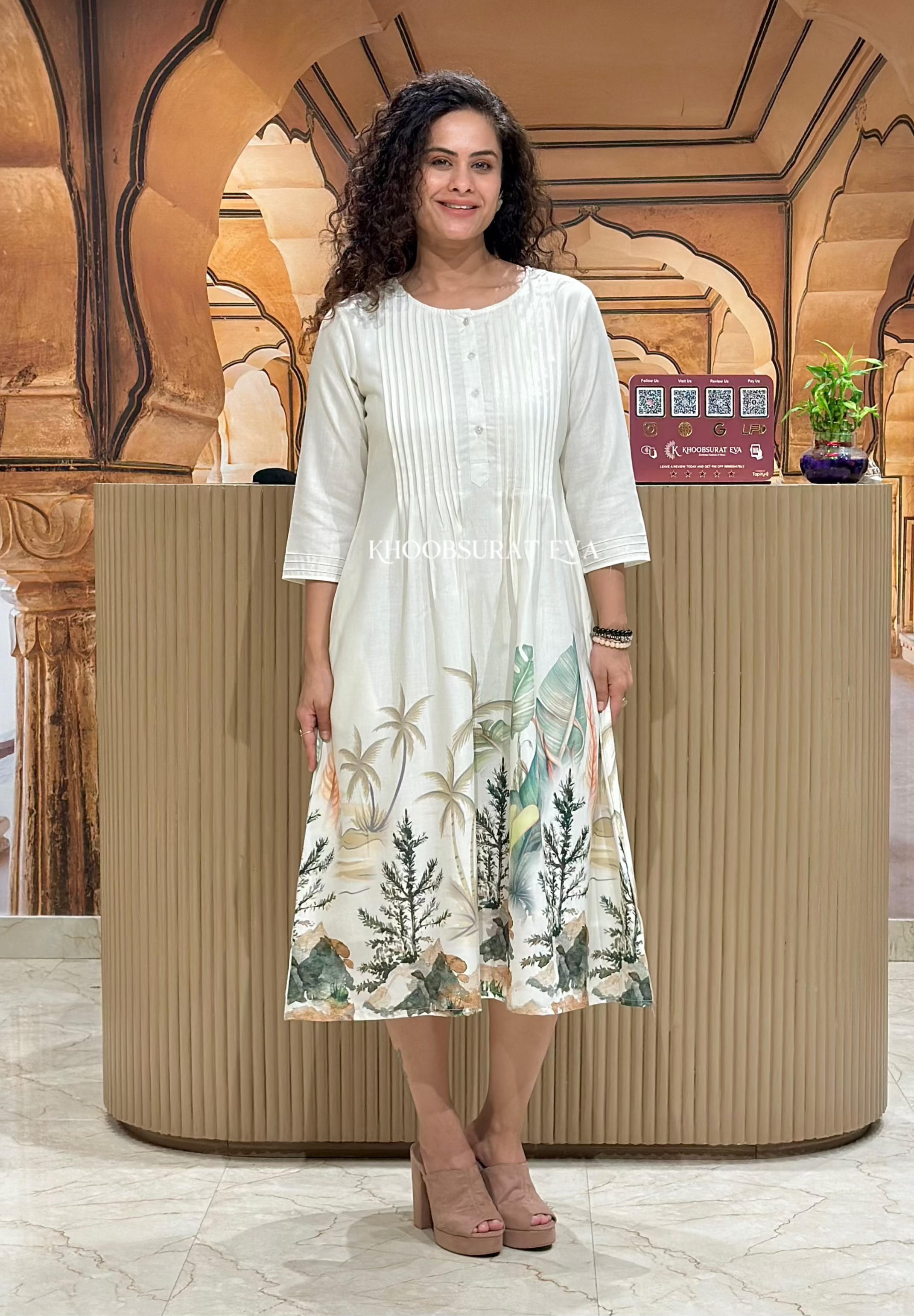 A woman standing in front of a neutral background wearing a white cotton knee-length A-line dress with 3/4 sleeves, featuring a floral botanical print at the hem and a collared neckline.