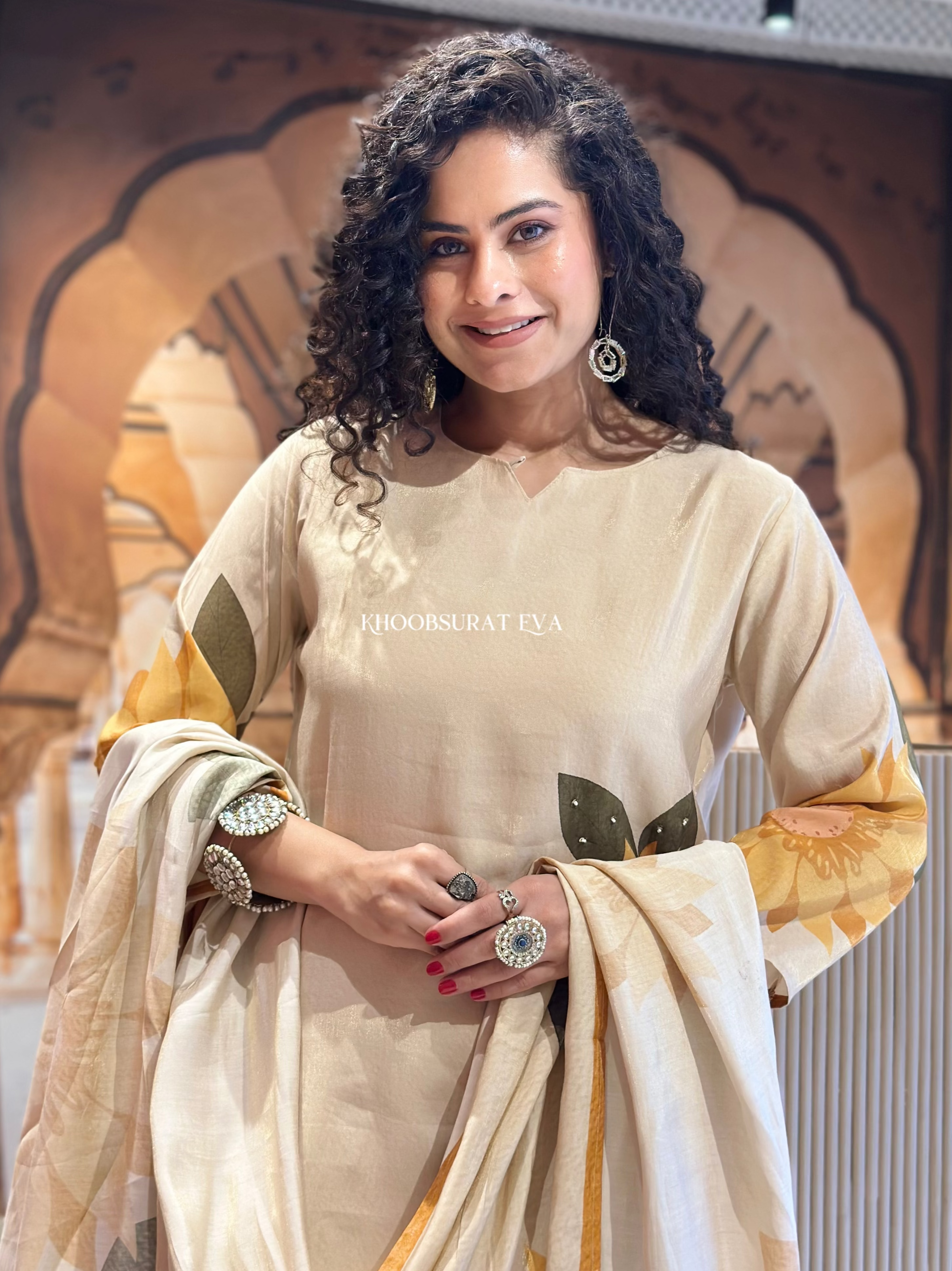 Woman wearing a beige suit with gold accents, standing against an ornate wall.